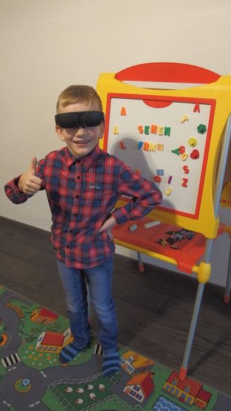 Kind trägt die NuEyes Pro und steht vor einer Tafel. Dabei streckt er den Daumen nach oben.
