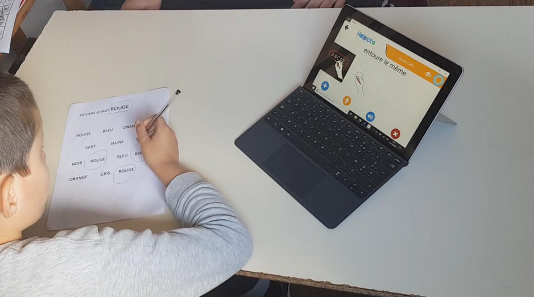 Kind sitzt mit Stift und Papier am Tisch. Das Tablet ist mit der Helpicto-App geöffnet.