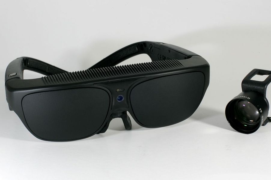Die NuEyes Pro ist dargestellt: Die schwarze Brille, Kamera und das Bluetooth-Steuergerät.