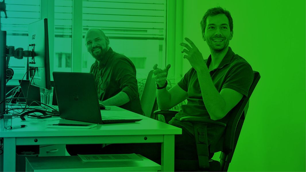 Zwei Männer sitzen an einem Schreibtisch mit Computer und Laptop. Das Bild ist in einem grünen Farbton.