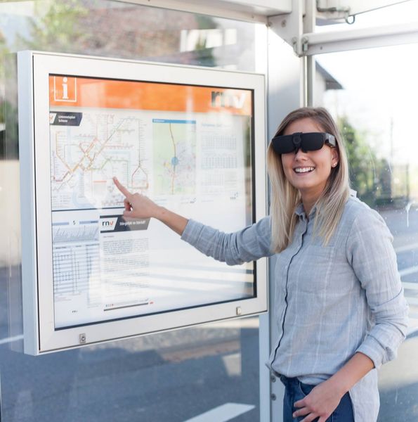 Eine Frau steht an der Bushaltestelle und zeigt auf einen Fahrplan. Dabei trägt sie die NuEyesPro.