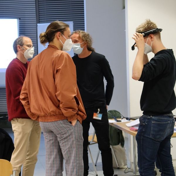 Vier Menschen stehen sich gegenüber. Einer hat eine VR Brille auf.