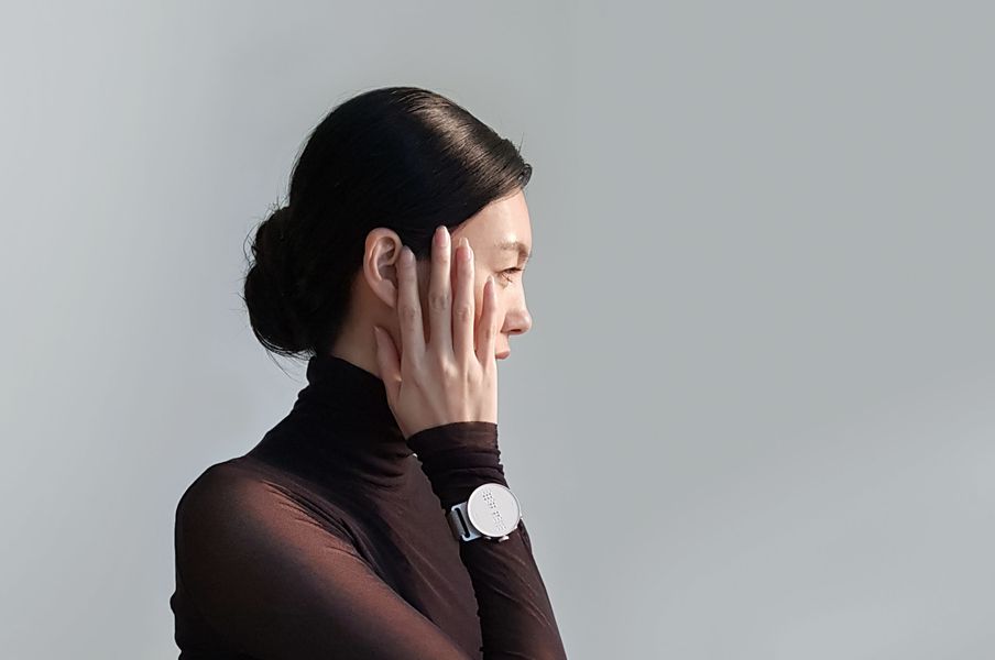 Eine schwarz gekleidete Frau ist von der Seite abgebildet. Sie fasst sich mit der rechten Hand ins Gesicht. An ihrem Handgelenkt ist die Dot Watch zu sehen.