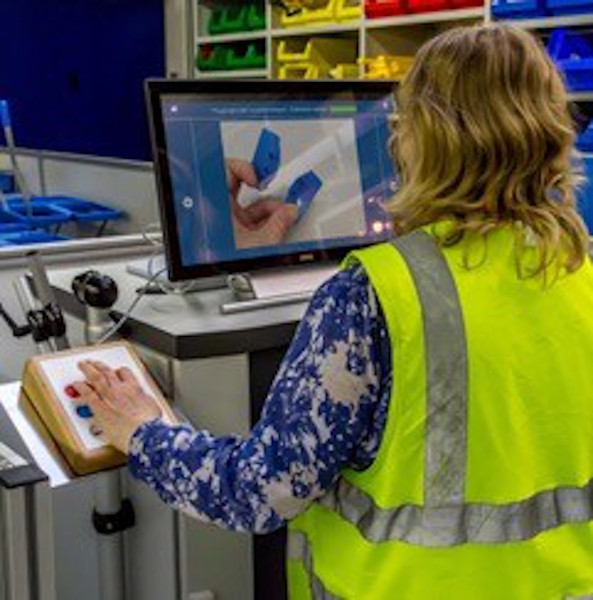 Eine Frau, die eine gelbe Warnweste trägt, bedient einen Computer mit einem Gerät, das sechs verschieden farbige Bedienungselemente hat.