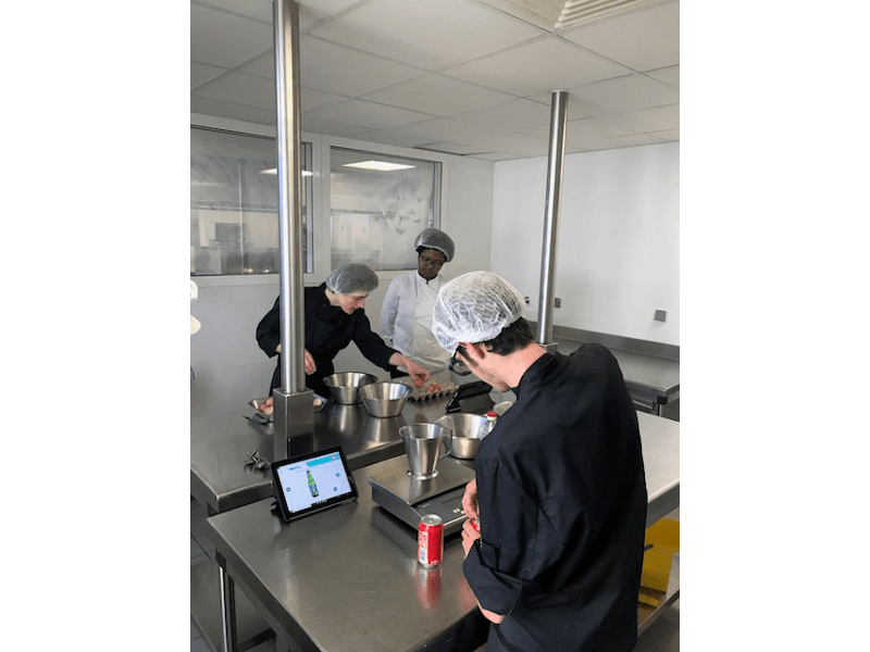 Drei Personen sind in einer Großküche. Ein Mann öffnet eine Dose und hat vor sich ein Tablet stehen, das die App Helpicto geöffnet hat. Zwei weitere Personen sind im Hintergrund zu sehen.