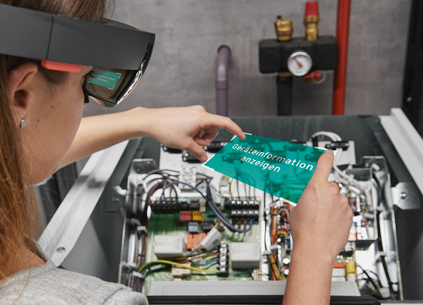 Eine Frau ist seitlich von hinten zu sehen. Sie trägt eine Datenbrille. Vor ihr ist ein offenes Gerät mit Kabeln. Sie hält ihre Hände darüber und ein artifizieller Bildschirm ist zwischen ihren Händen eingeblendet. Darauf steht: Information anzeigen.