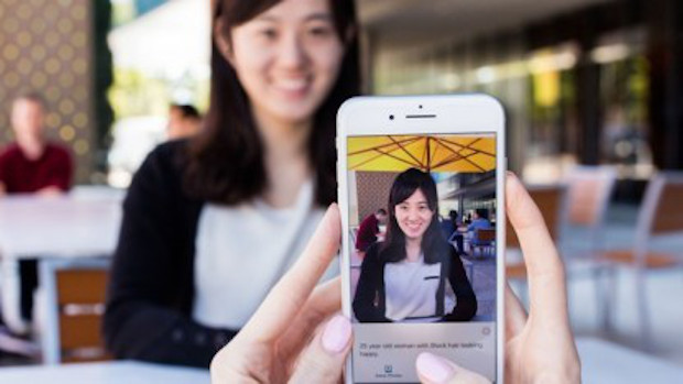Eine Hand hält ein Iphone auf welchem die App Seeing AI geöffnet ist. Mit Hilfe der Iphone-Kamera wird eine Frau auf dem Display erkannt, welche im Hintergrund des Bildes unscharf zu erkennen ist.