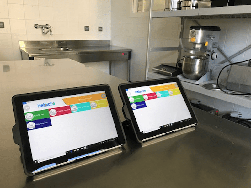 Zwei Tablets stehen auf einer Küchenplatte in einer Großküche. Auf beiden Bildschirmen ist die Helpicto App geöffnet.