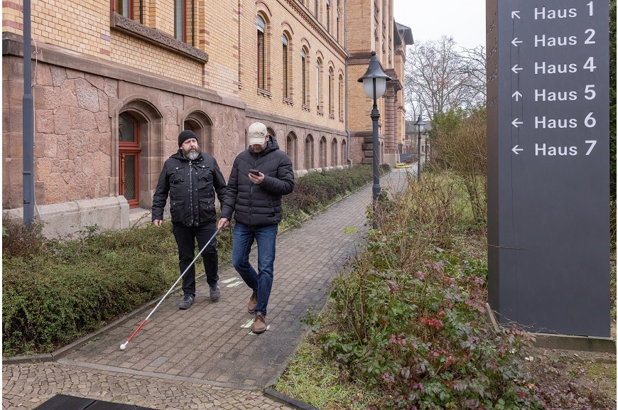 Zwei Personen gehen auf einem Gehweg. Die eine Person hat ein Smartphone in der einen Hand, und in der anderen einen Langstock.