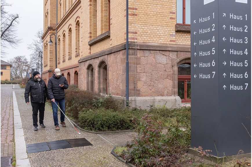 Zwei Personen gehen auf einem Gehweg. Die eine Person hat ein Smartphone in der einen Hand, und in der anderen einen Langstock.