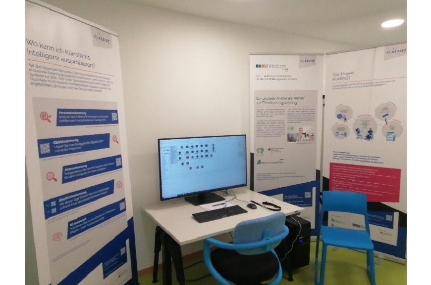 Ein Computerbildschirm auf einem Schreibtisch. Neben dem Computer sind Plakate mit Informationen